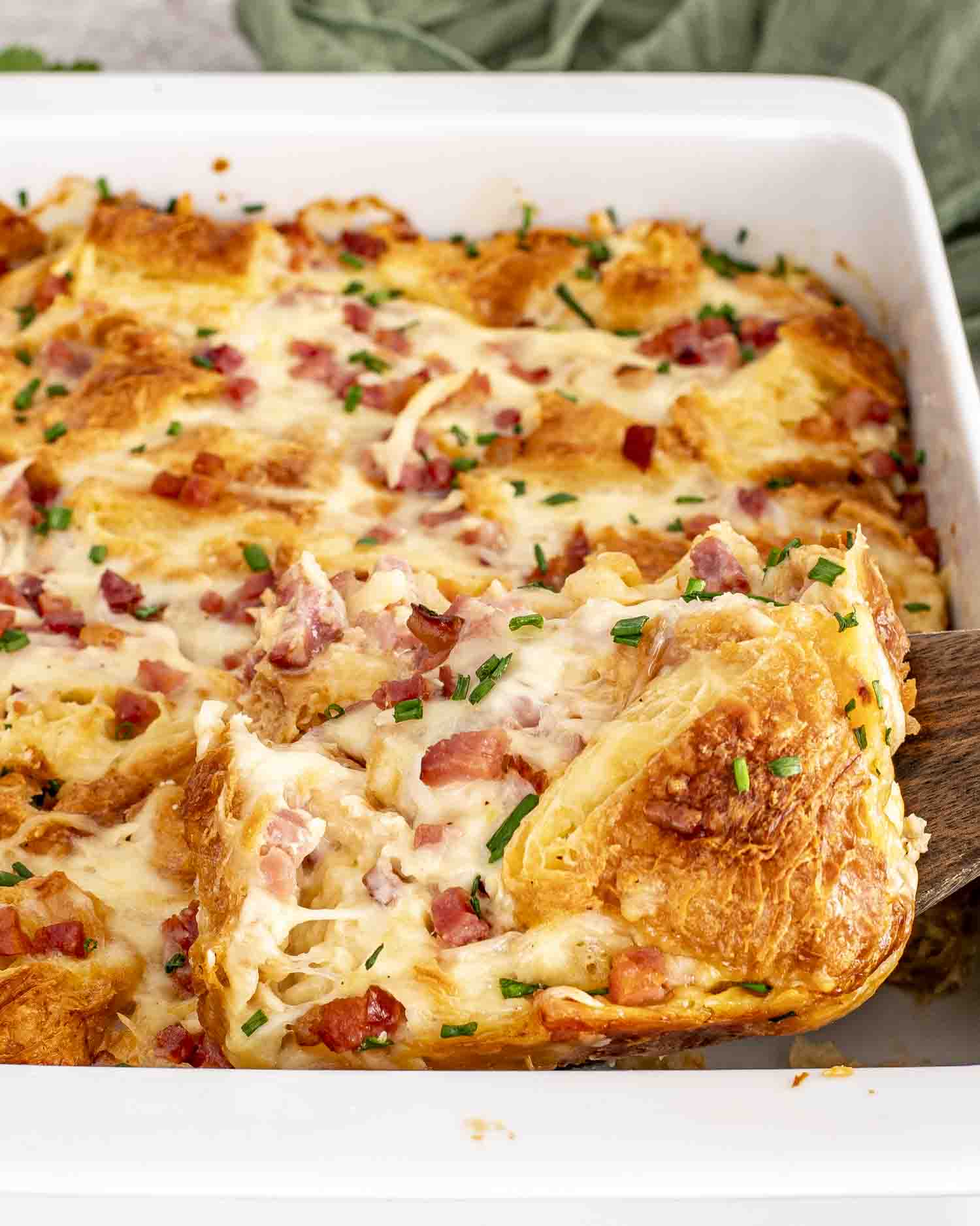 Cheesy croissant egg bake scooped from baking dish, layers of ham and croissants visible, chive garnish.