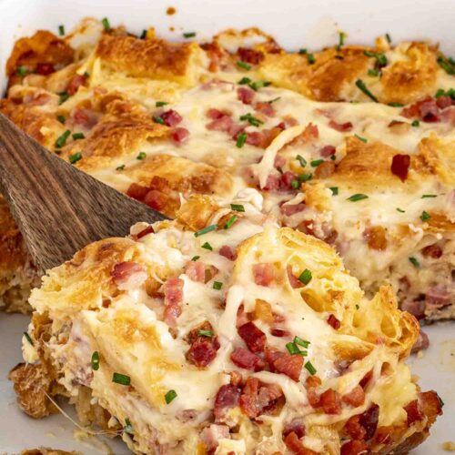 Baking dish with ham and cheese croissant egg bake, wooden spatula lifting golden cheesy portion out.