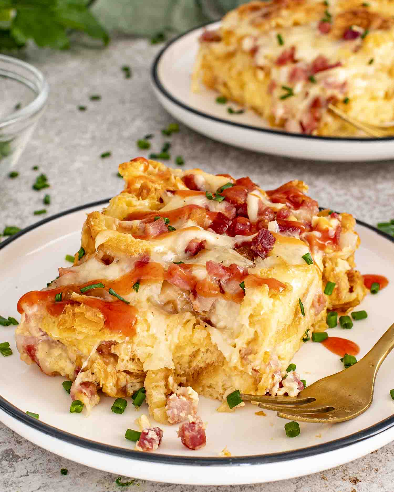 Close-up slice of ham and cheese croissant egg bake on white plate, garnished with chives and drizzle.