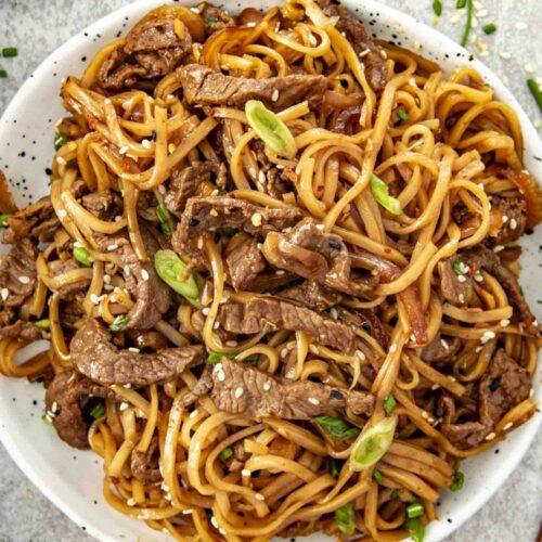 Overhead view of Mongolian beef noodles on a white plate, garnished with scallions and sesame seeds.