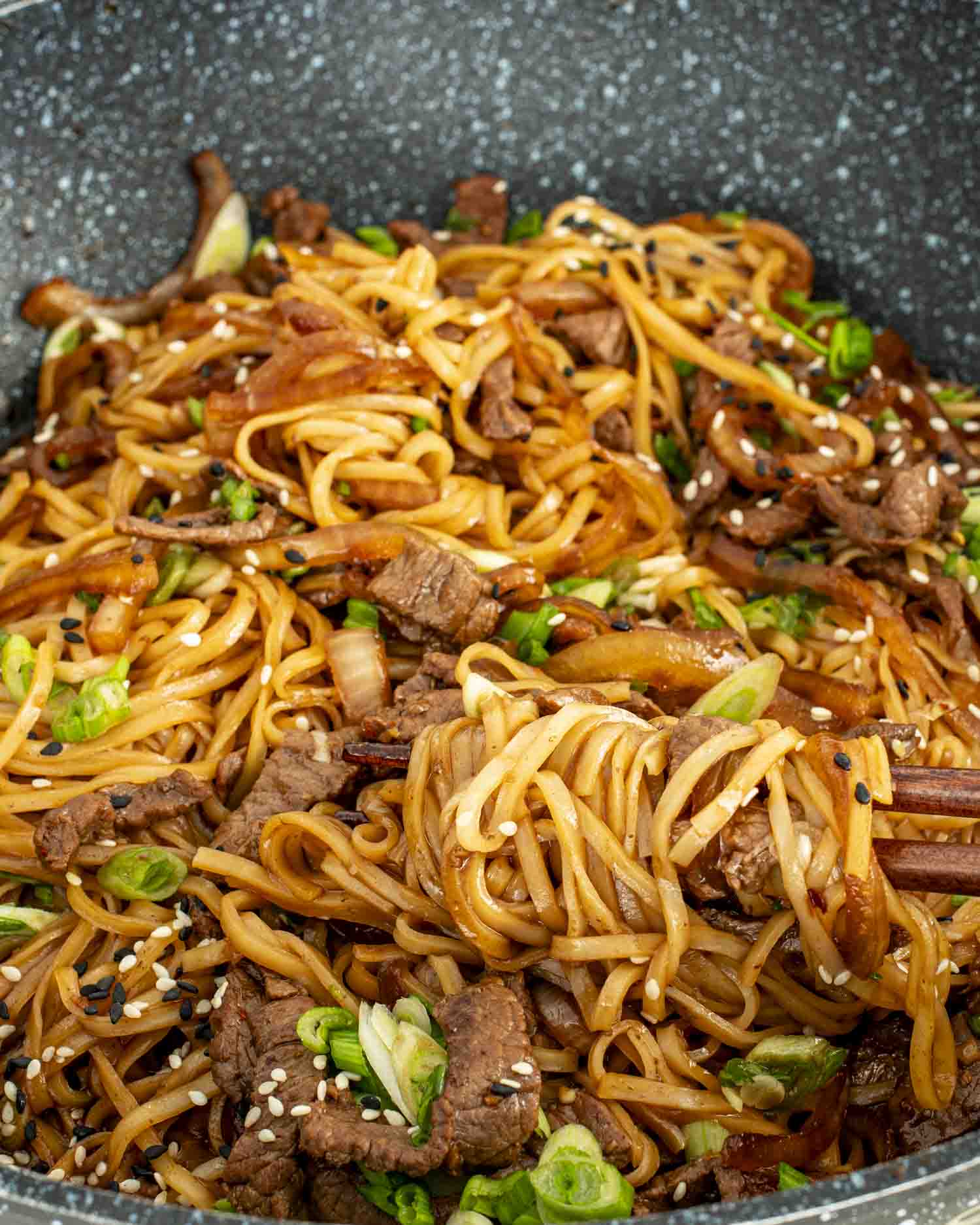 Close‑up of wok‑fried Mongolian beef noodles tossed with onions, sesame seeds, and tender strips of beef.