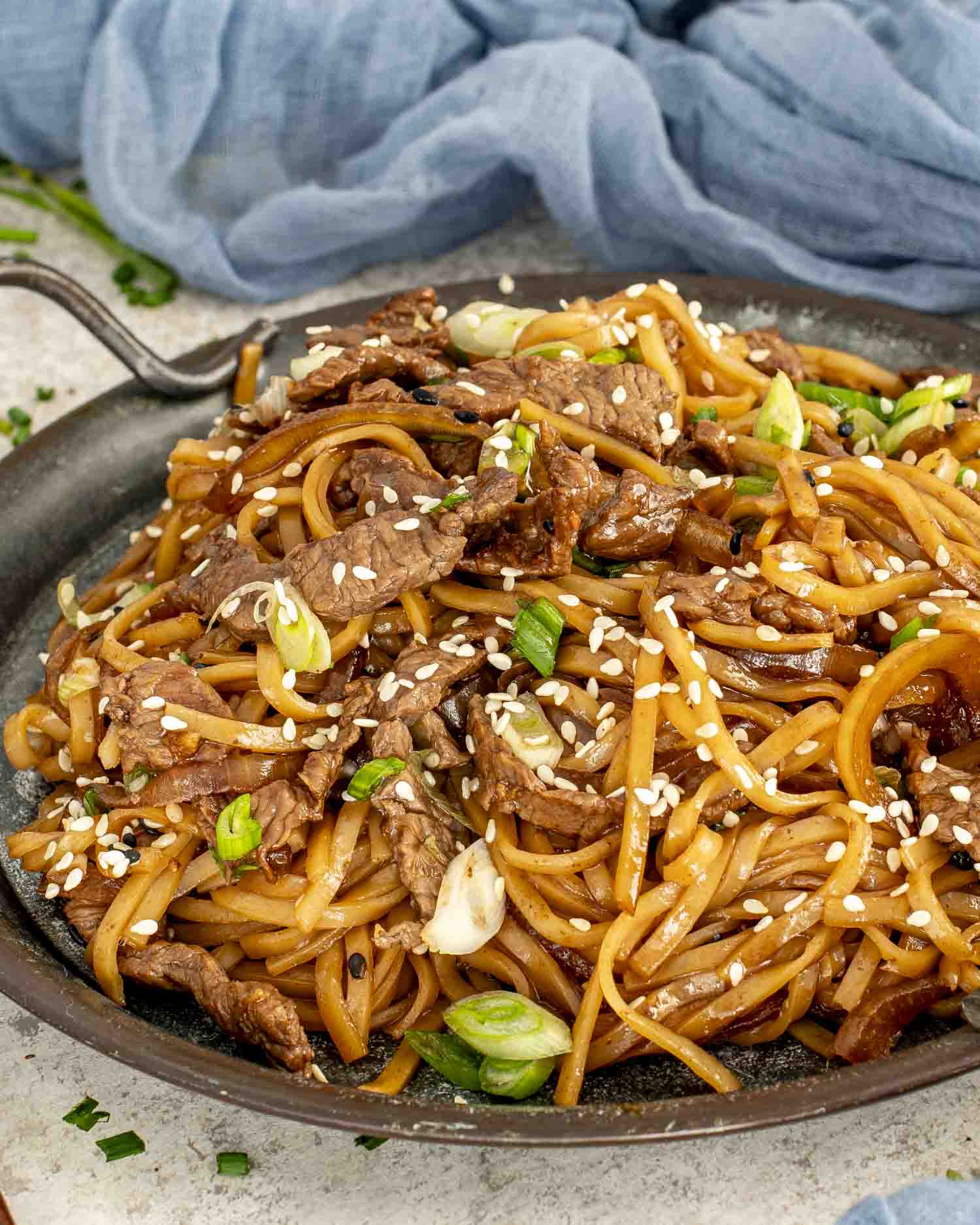 A rustic pan filled with glossy Mongolian beef noodles, sprinkled with sesame seeds and green onions.