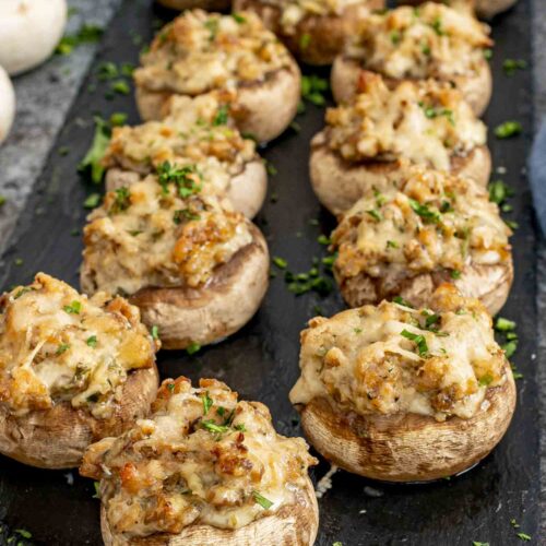 Row of sausage stuffed mushrooms on dark slate board, topped with cheese, parsley, perfect for party appetizer platter.
