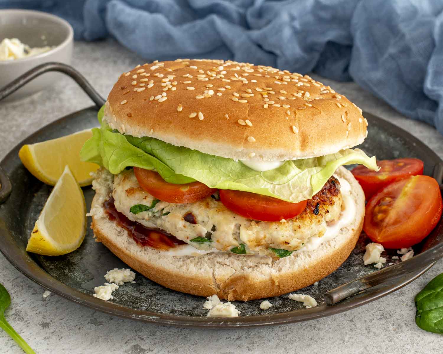 Spinach and feta chicken patty sandwich with lettuce and tomato on a sesame bun, served with lemon wedges.