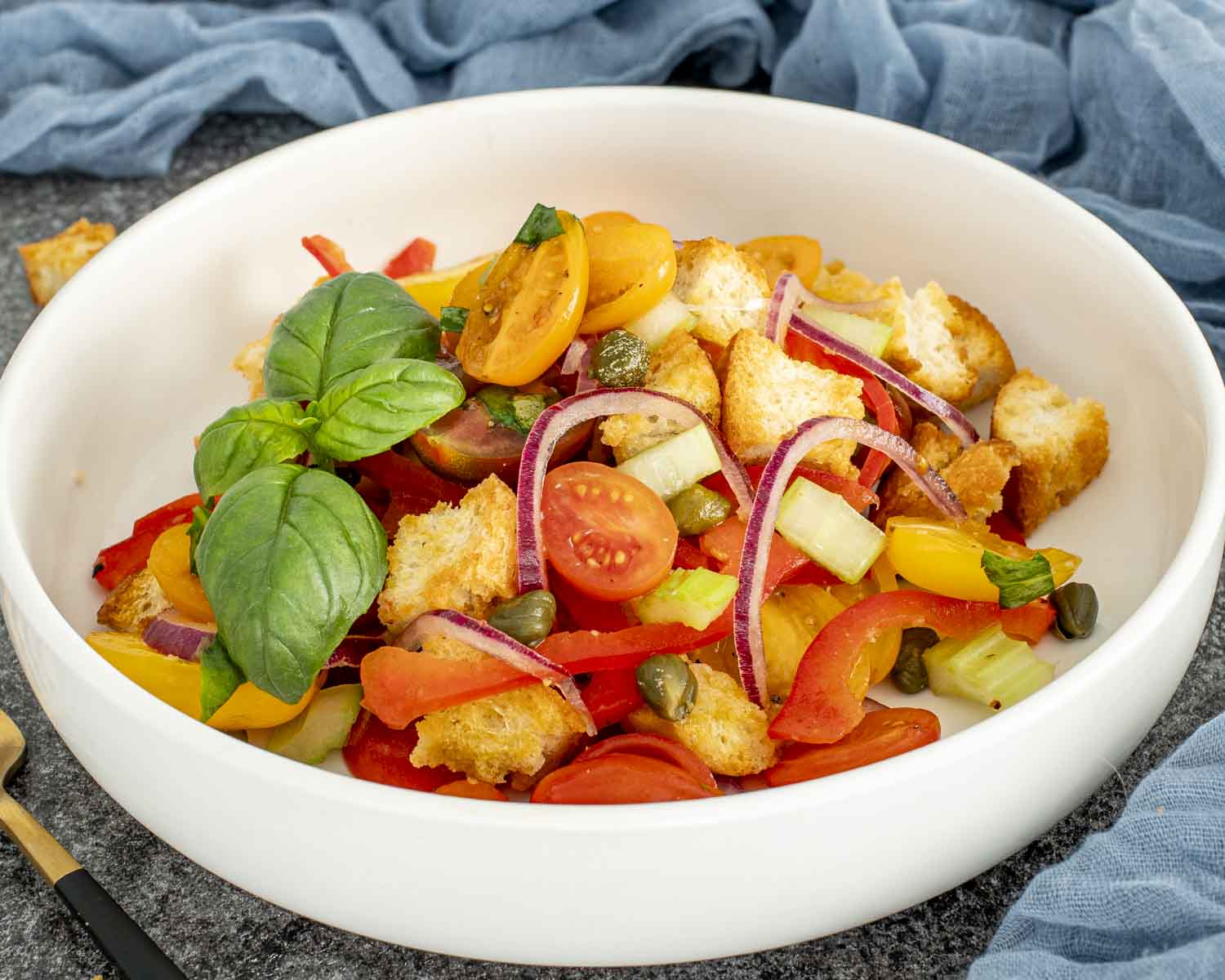 Panzanella salad being tossed with wooden spoon, featuring crusty bread, tomatoes, bell peppers, capers, and fresh basil.