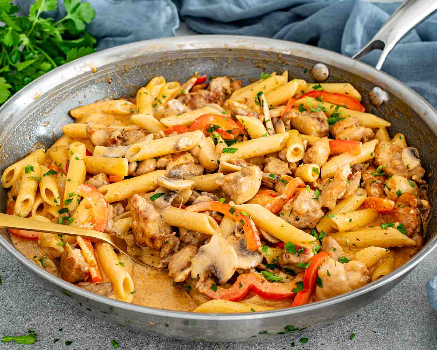 Creamy Cajun chicken pasta cooking in a wide skillet with penne, chicken pieces, mushrooms, and fresh parsley garnish.