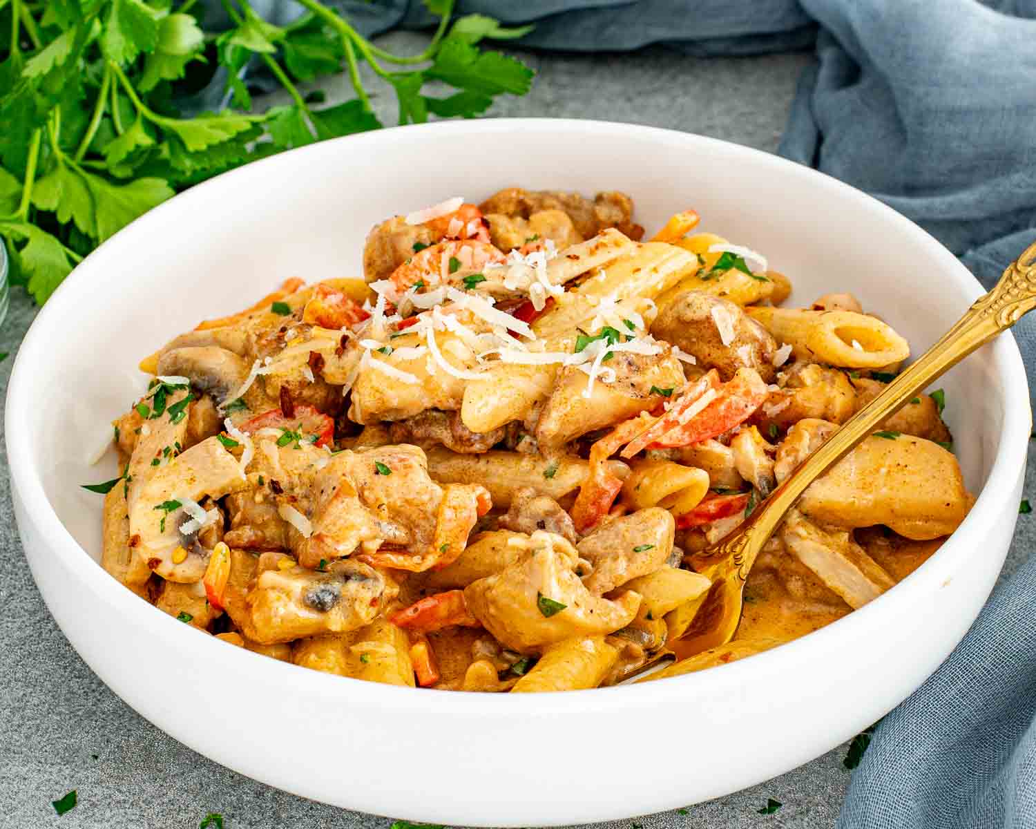 Close up of creamy Cajun chicken pasta in a white bowl with mushrooms, peppers, and a golden spoon.
