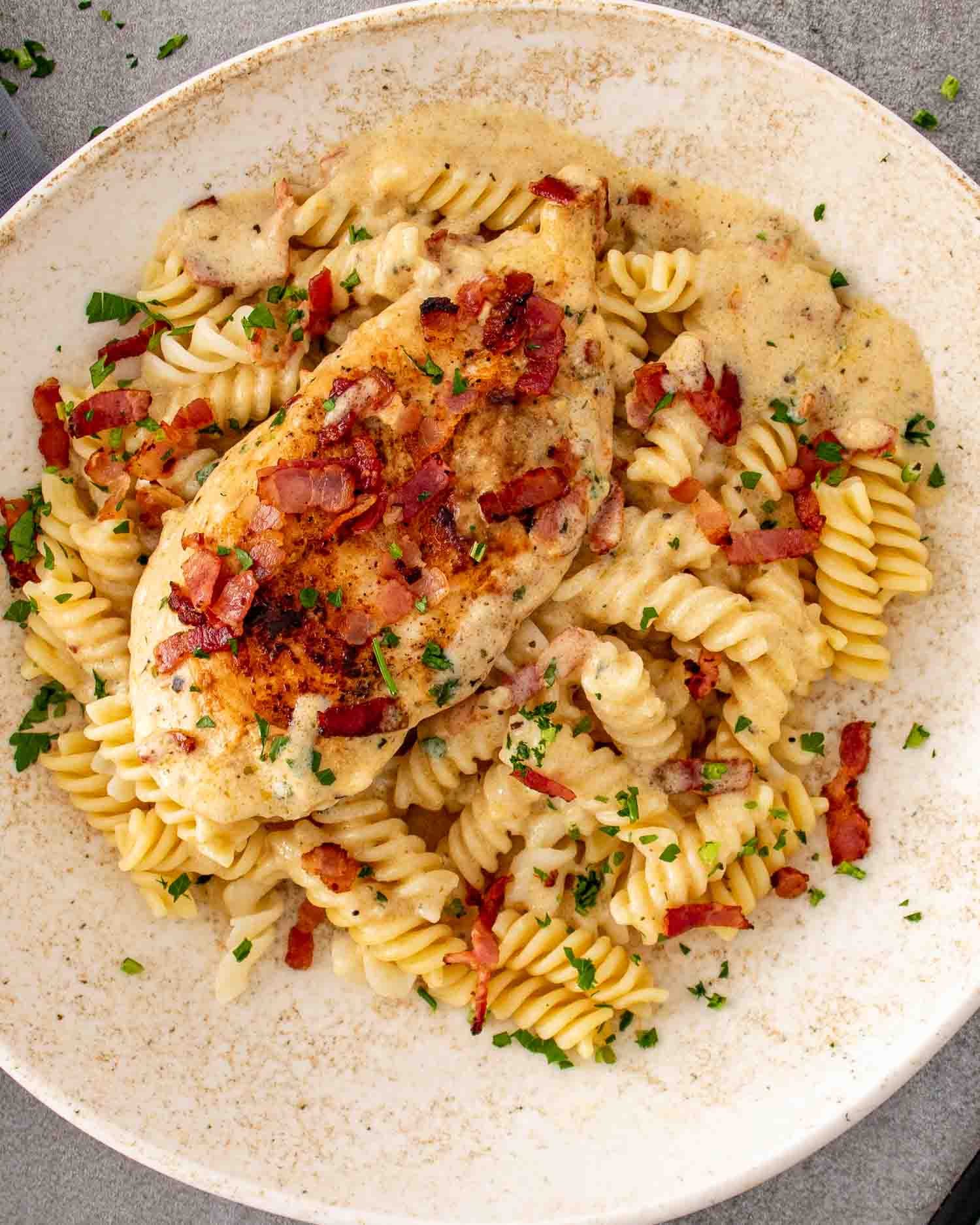 Plated creamy garlic parmesan chicken served over pasta with bacon, parsley, and extra sauce spooned on top.