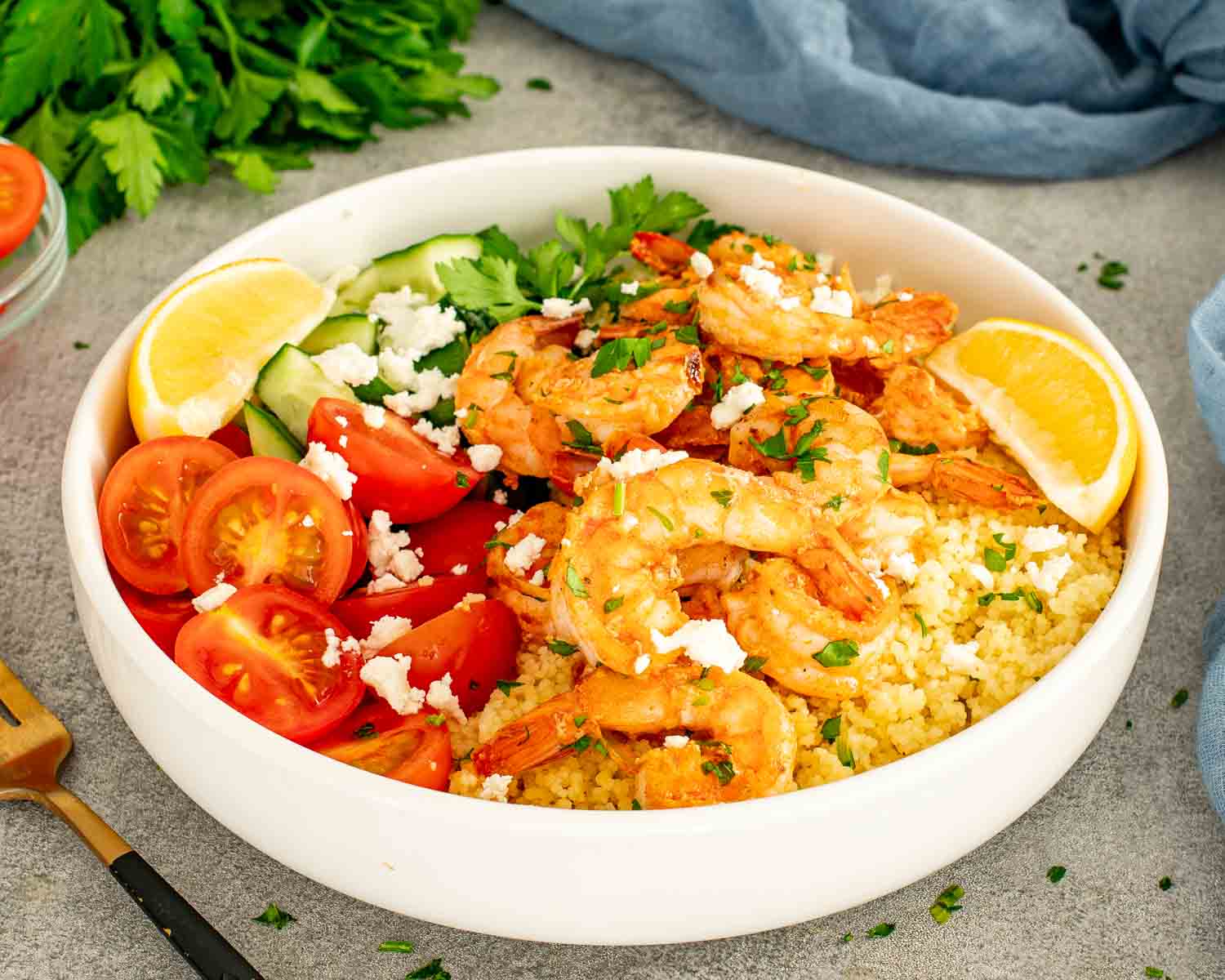 Fresh garlic shrimp couscous bowl with vibrant tomatoes, cucumber, feta cheese, and parsley on gray surface.