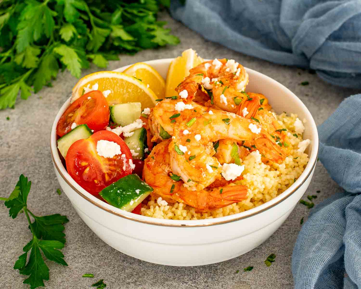Garlic shrimp couscous bowl with lemon wedges, cherry tomatoes, cucumber, and feta in white serving bowl.