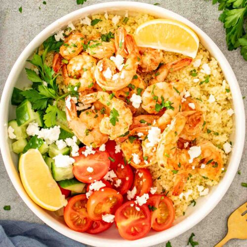 Garlic shrimp couscous bowl topped with cherry tomatoes, cucumber, feta, fresh parsley, and lemon wedges on light background.
