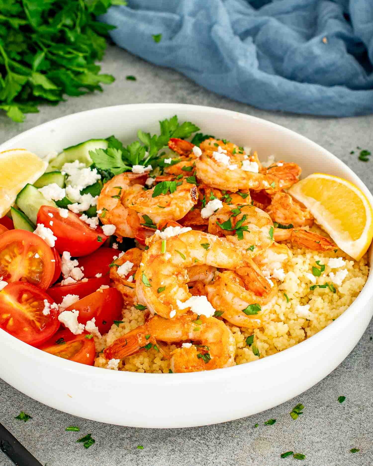 Garlic shrimp couscous bowl topped with cherry tomatoes, cucumber, feta, fresh parsley, and lemon wedges on light background.