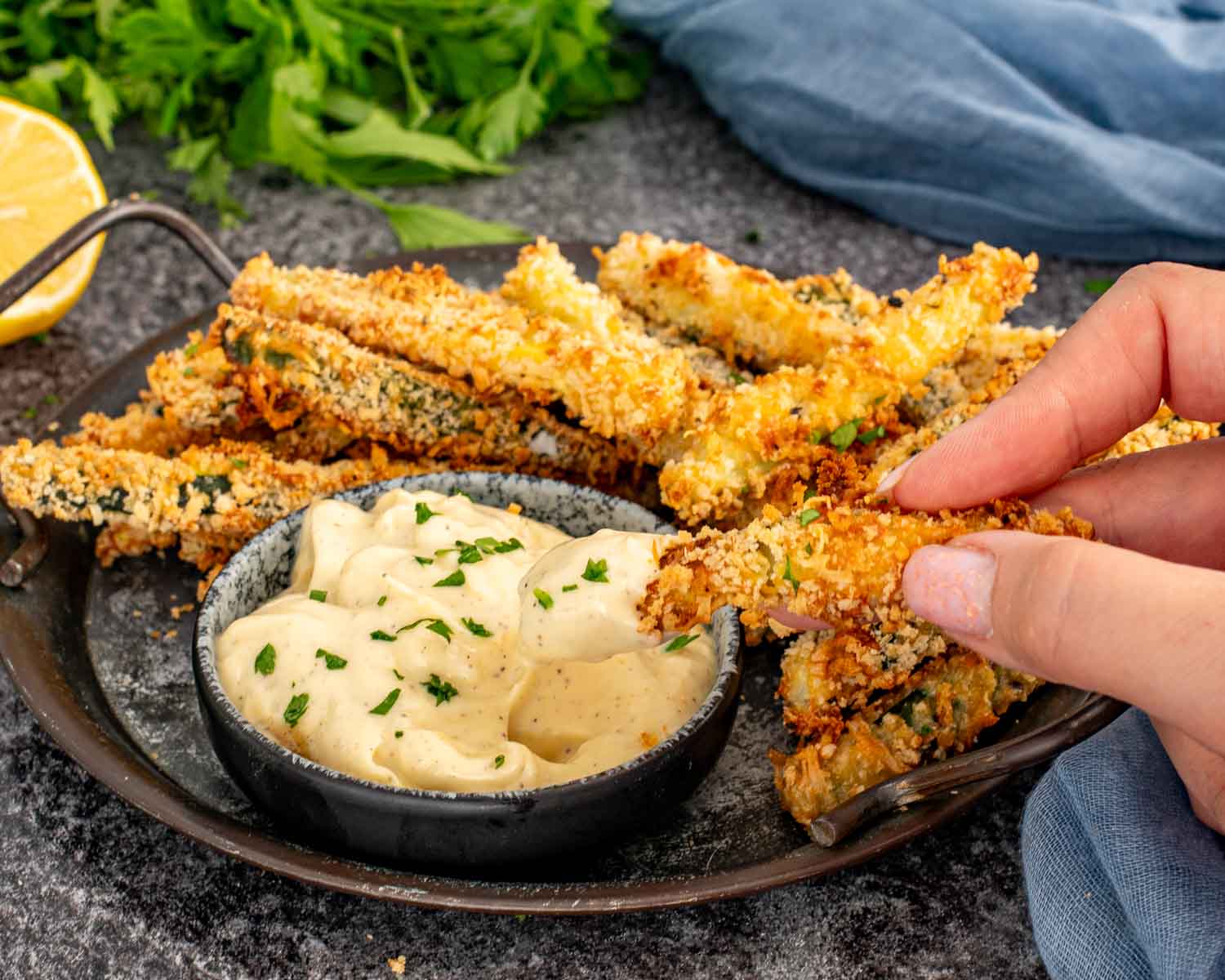 Hand dipping crispy zucchini fry into creamy garlic aioli sauce on serving tray.