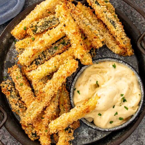 Golden baked zucchini fries piled on a dark tray served with homemade garlic aioli dipping sauce.