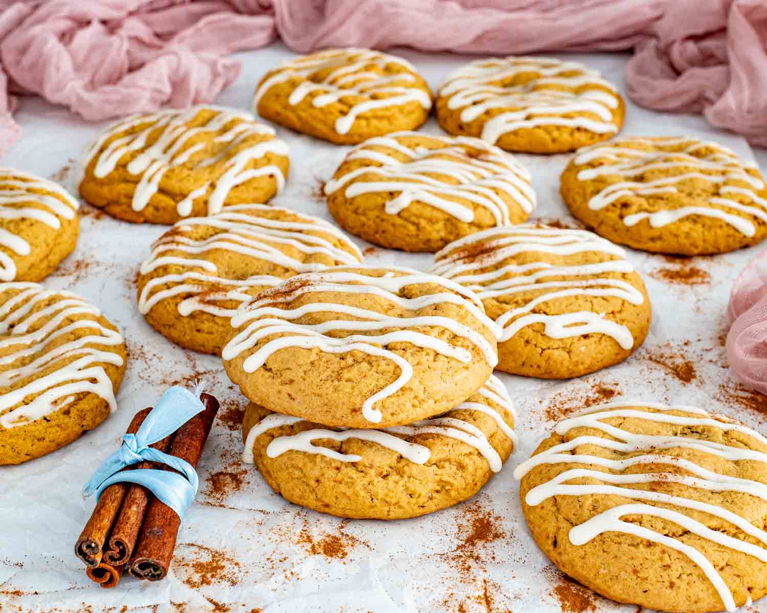 Fresh batch of cinnamon roll cookies with cream cheese glaze spread across parchment with cinnamon sticks and soft textured background.