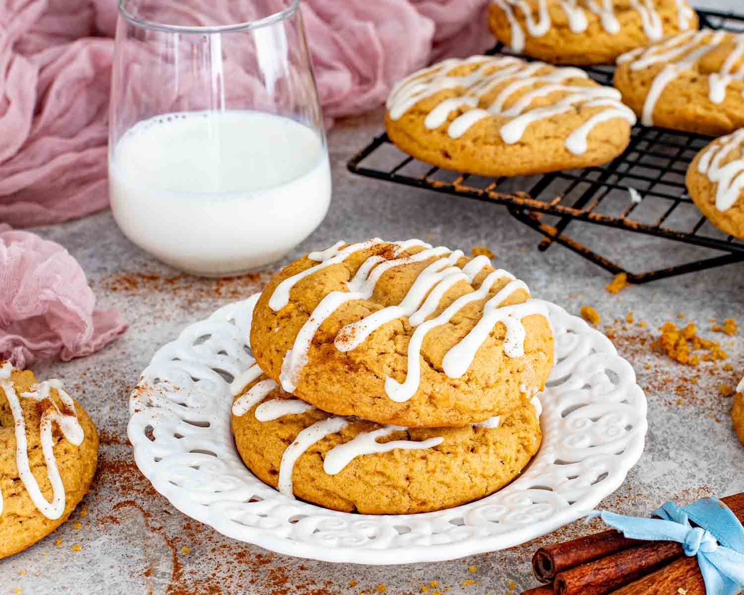 Cinnamon roll cookies on plate with cream cheese glaze, glass of milk, and cooling rack of cookies in background.