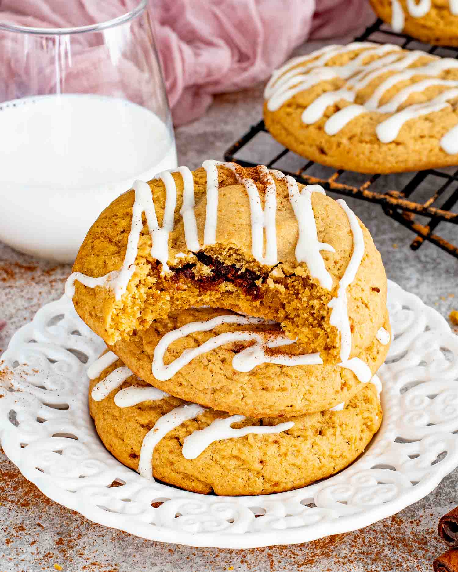 Stacked cinnamon roll cookies with cream cheese glaze on decorative plate, one bitten showing gooey cinnamon center with glass of milk.