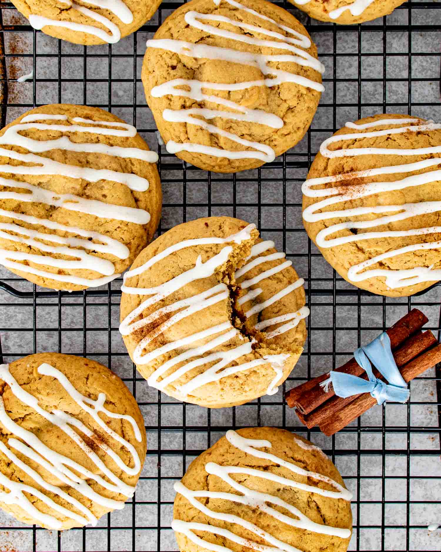 Cinnamon roll cookies cooling on a wire rack topped with cream cheese glaze and dusted with cinnamon beside tied cinnamon sticks.
