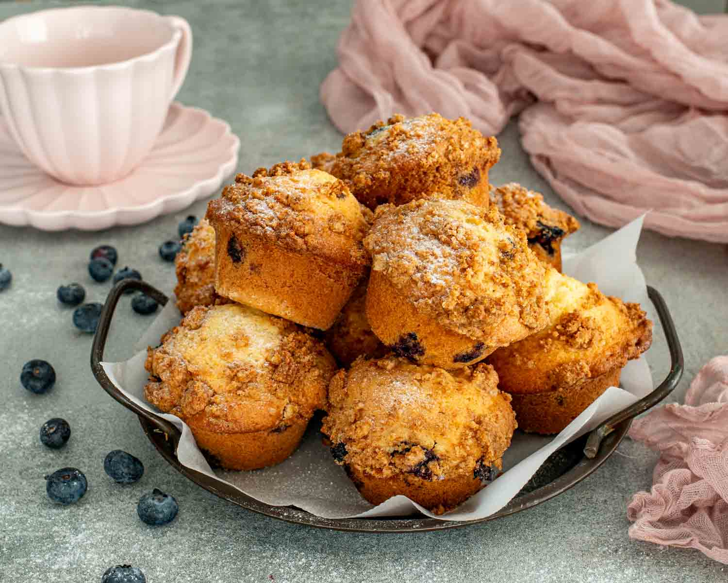 Jumbo blueberry crumb muffins stacked in tray lined with parchment paper, golden crumb topping and tall muffin tops.