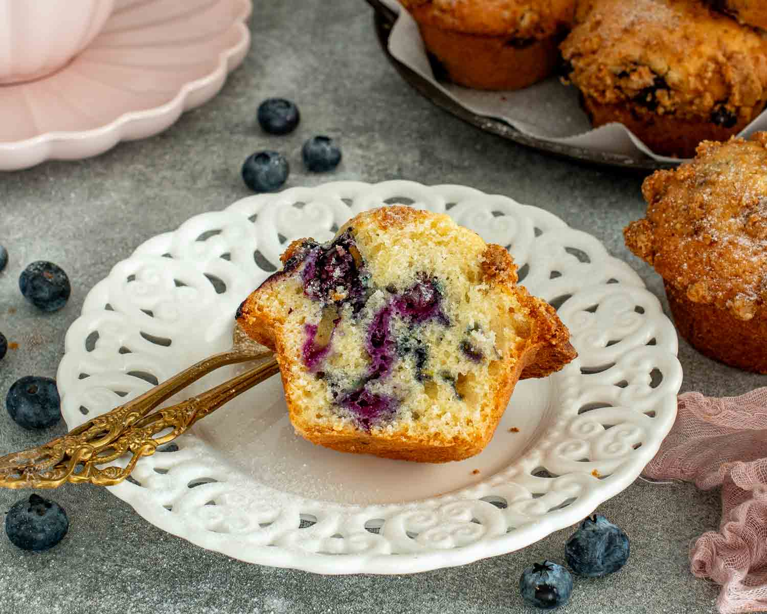 Bakery style blueberry crumb muffins on tray with pink cup and plate, styled cozy kitchen scene with linen cloth.