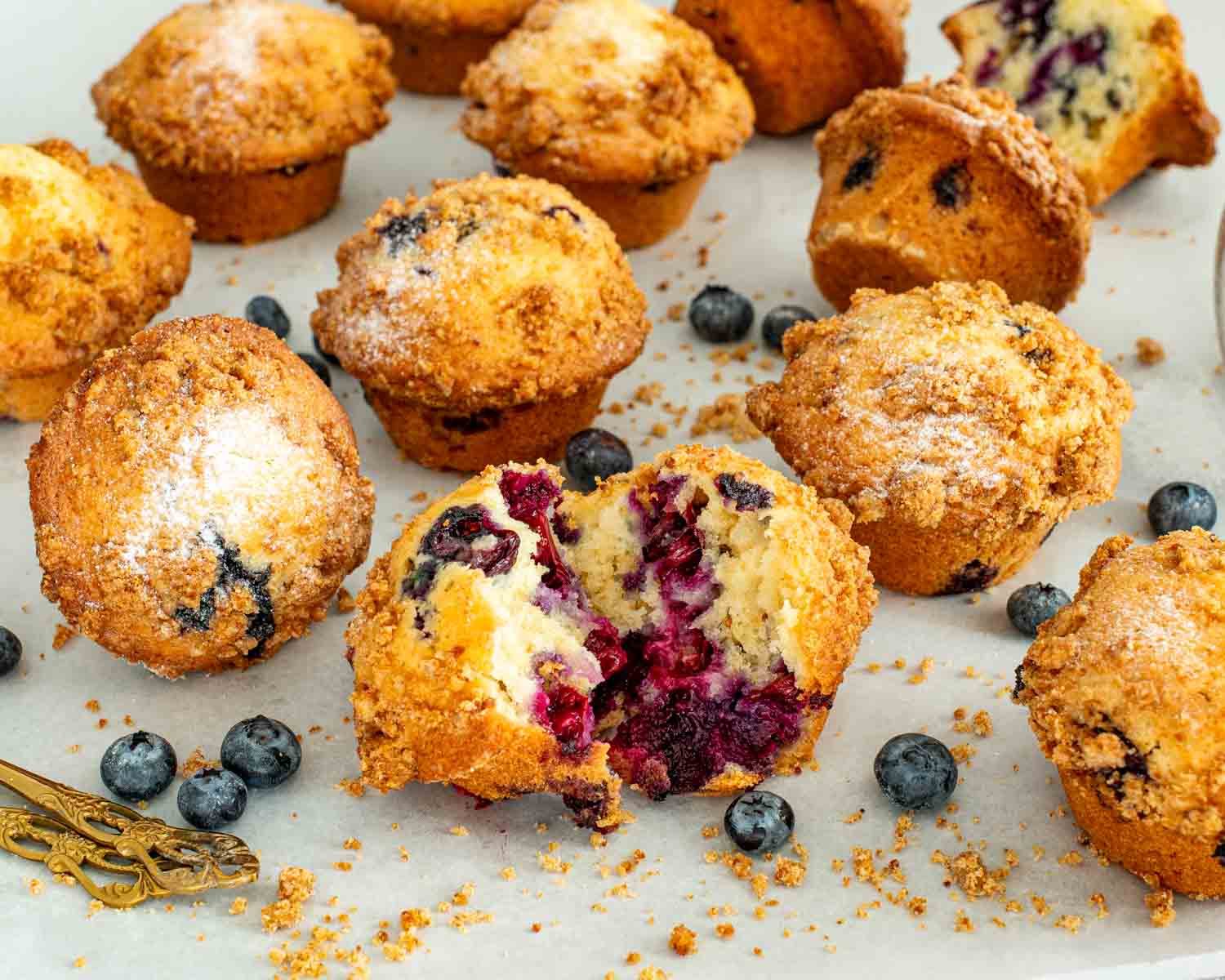 Blueberry crumb muffins on parchment paper with scattered crumbs and fresh blueberries around, bakery style homemade muffins.