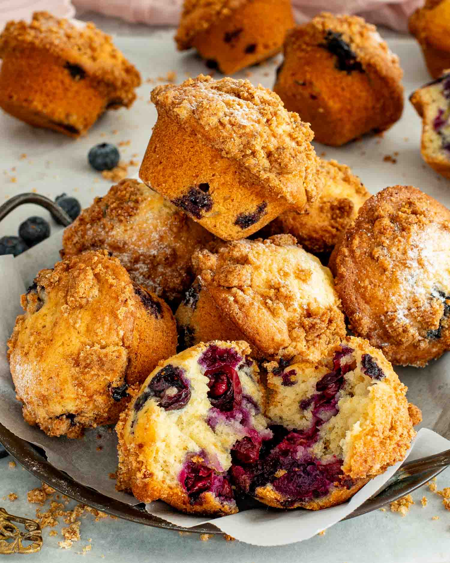 Stacked jumbo blueberry crumb muffins on parchment lined tray with powdered sugar and fresh blueberries on gray surface.