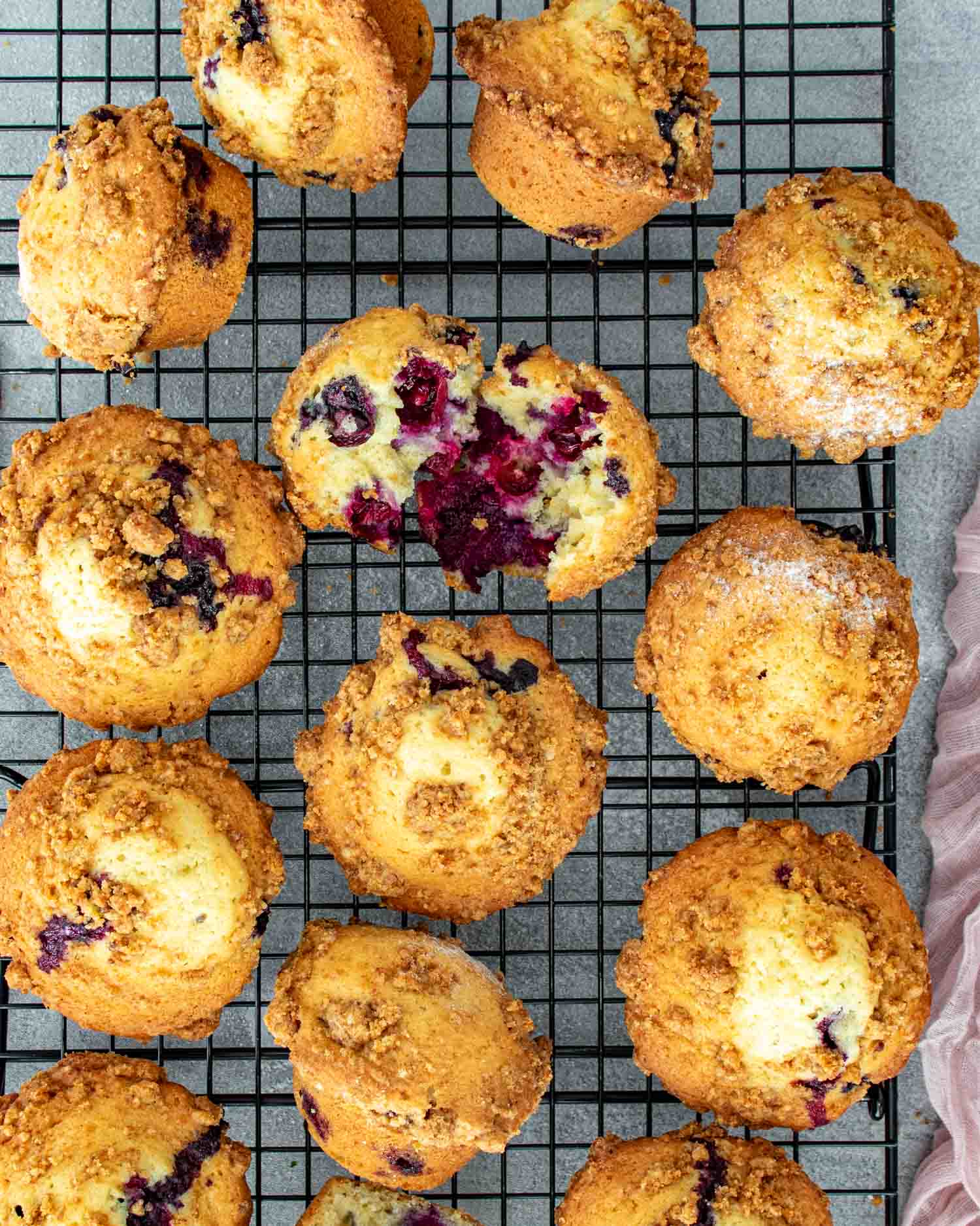 Overhead view of jumbo blueberry crumb muffins on cooling rack with golden crumb topping and one muffin broken open.