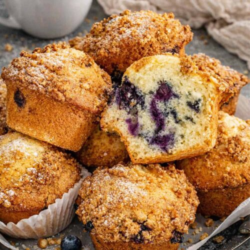 Close up of jumbo blueberry crumb muffins stacked on parchment lined tray with crumb topping and blueberries visible inside.