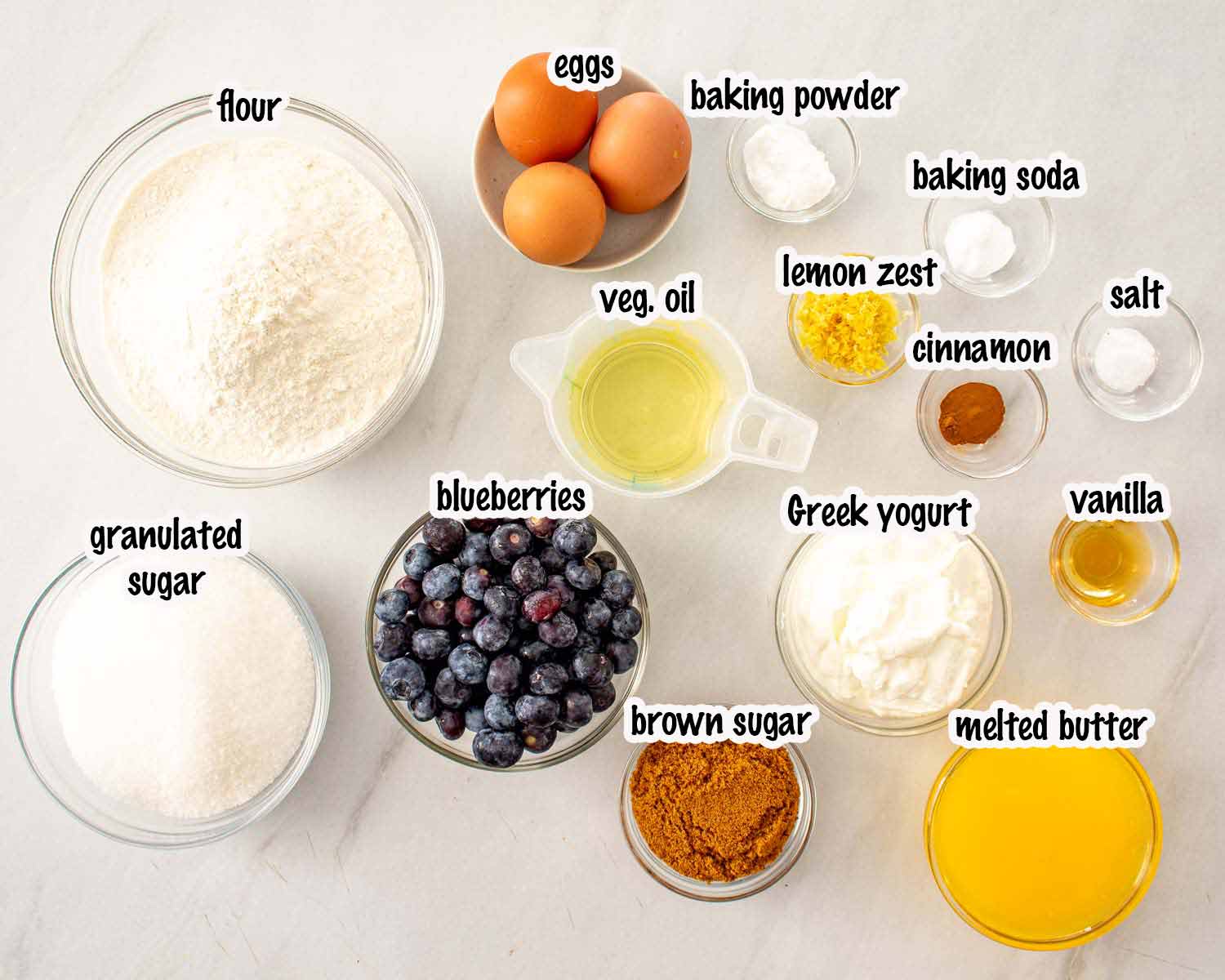 Overhead view of baking ingredients in glass bowls including flour, blueberries, sugar, yogurt, eggs, butter, oil, and spices.