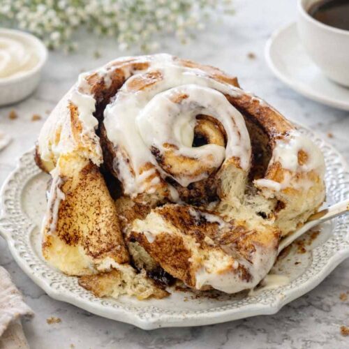 A frosted jumbo cinnamon roll served on a plate next to a cup of black coffee and fresh cinnamon sticks.