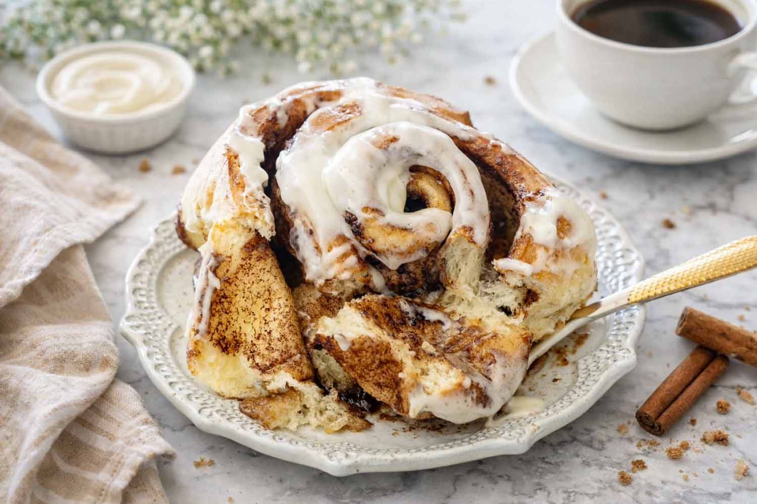 A frosted jumbo cinnamon roll served on a plate next to a cup of black coffee and fresh cinnamon sticks.