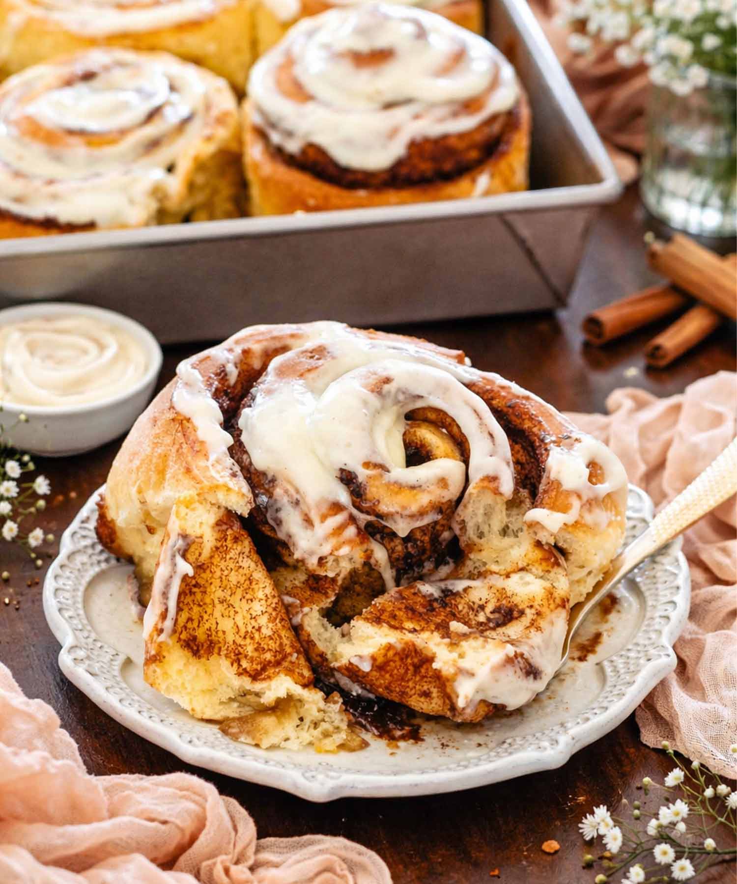Close up of a frosted cinnamon roll on a decorative plate with a gold fork and a pan in background.