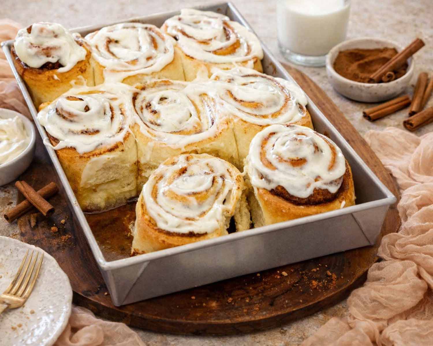 Side view of a baking pan filled with fluffy cinnamon rolls topped with melting cream cheese icing and cinnamon sticks.