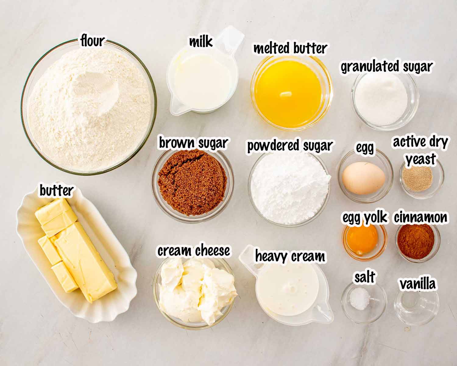 An overhead view of various ingredients in glass bowls labeled with text including flour sugar butter cream cheese and yeast. These ingredients are needed to make cinnamon rolls.