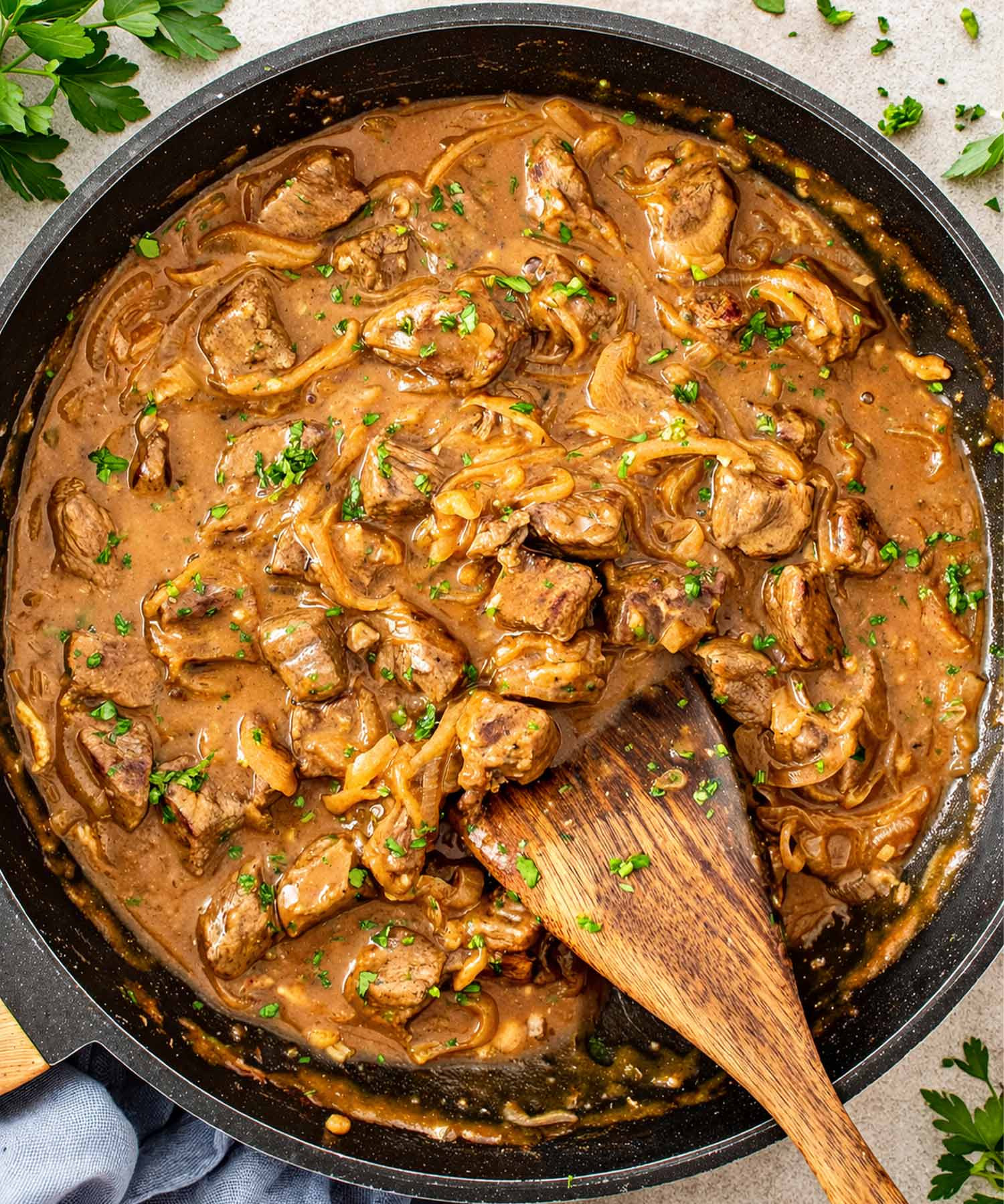 Overhead view of beef and onion skillet with browned beef, soft onions, and rich gravy being stirred with a wooden spoon.