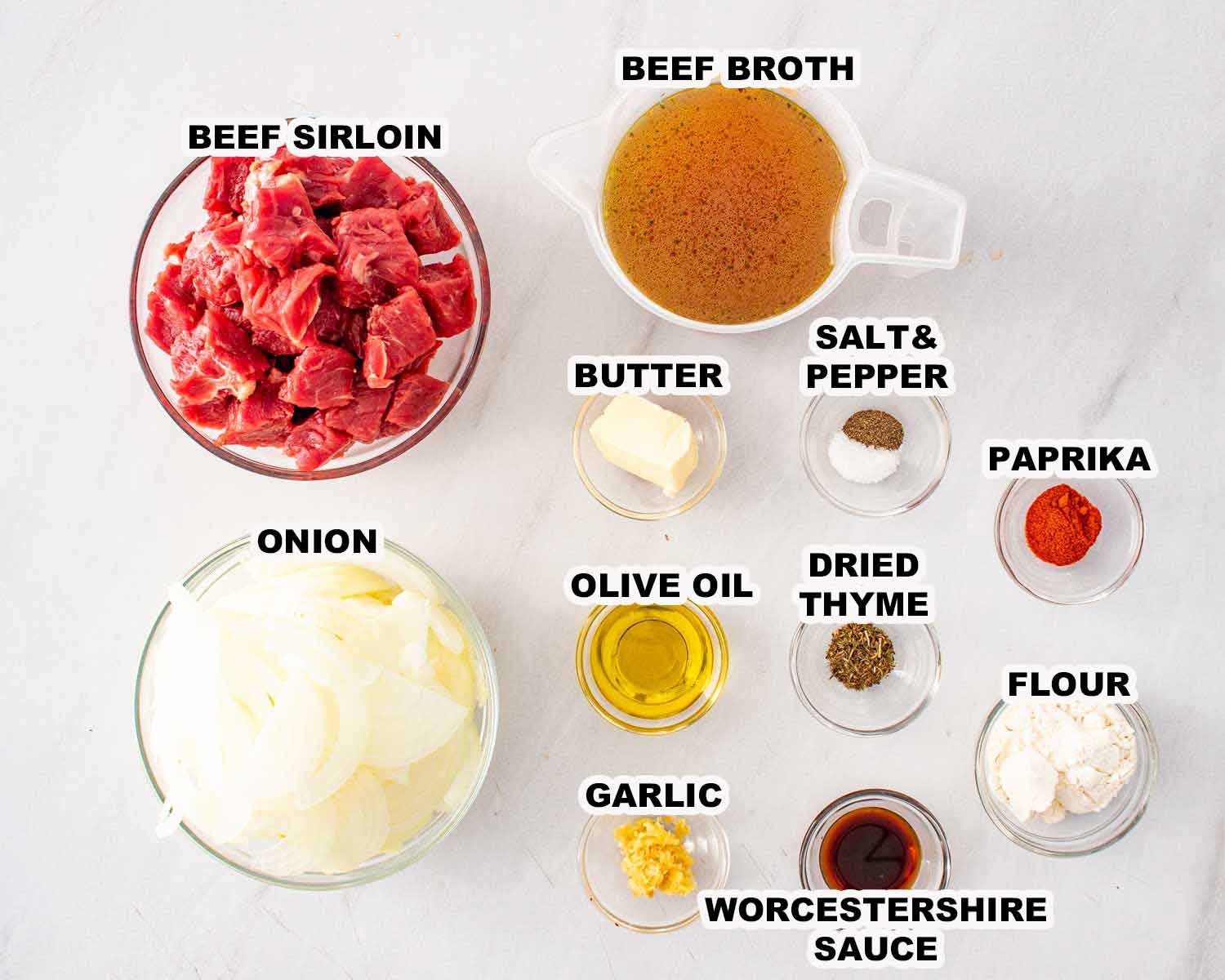 Ingredients for beef and onion skillet including beef sirloin, onions, garlic, broth, Worcestershire sauce, flour, butter, and spices arranged on table.