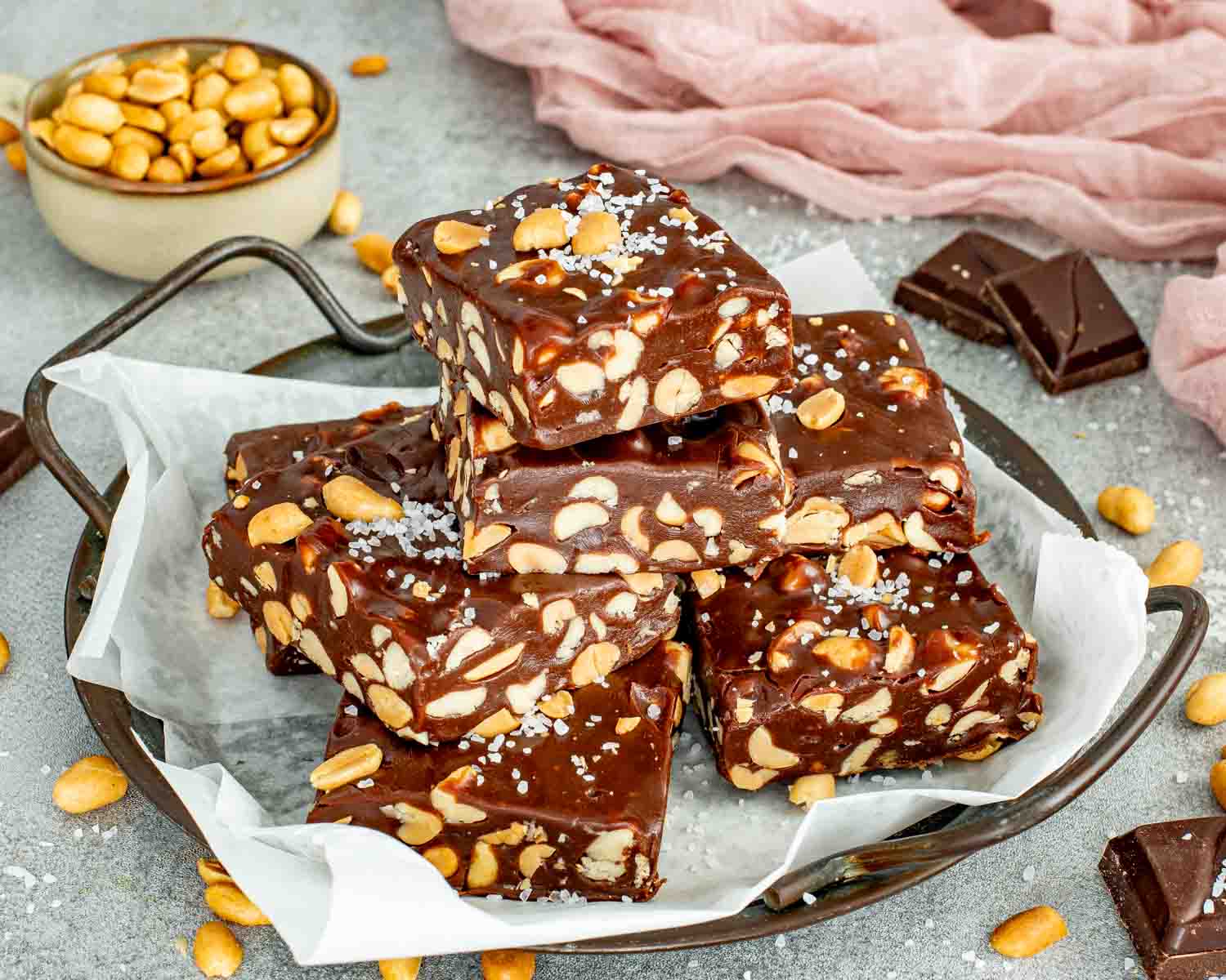 A pile of chewy chocolate peanut bars on parchment paper inside a rustic metal tray with salt.