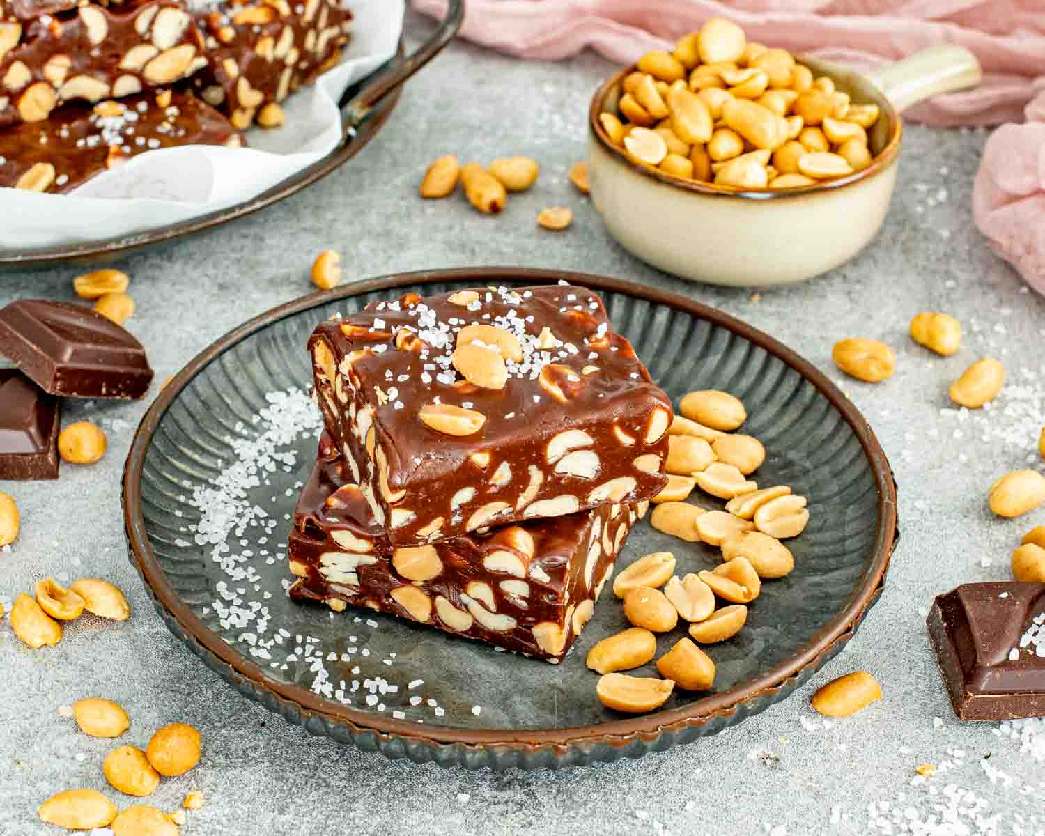 Square chocolate peanut bars sprinkled with flaky sea salt on a dark metal plate with loose peanuts.