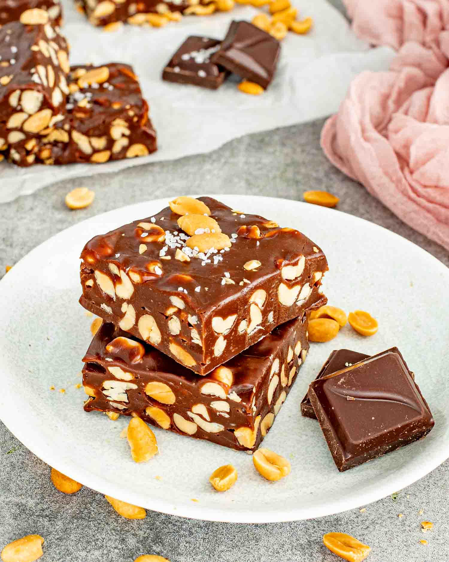 Two thick chocolate peanut bars stacked on a white plate next to dark chocolate chunks and peanuts.