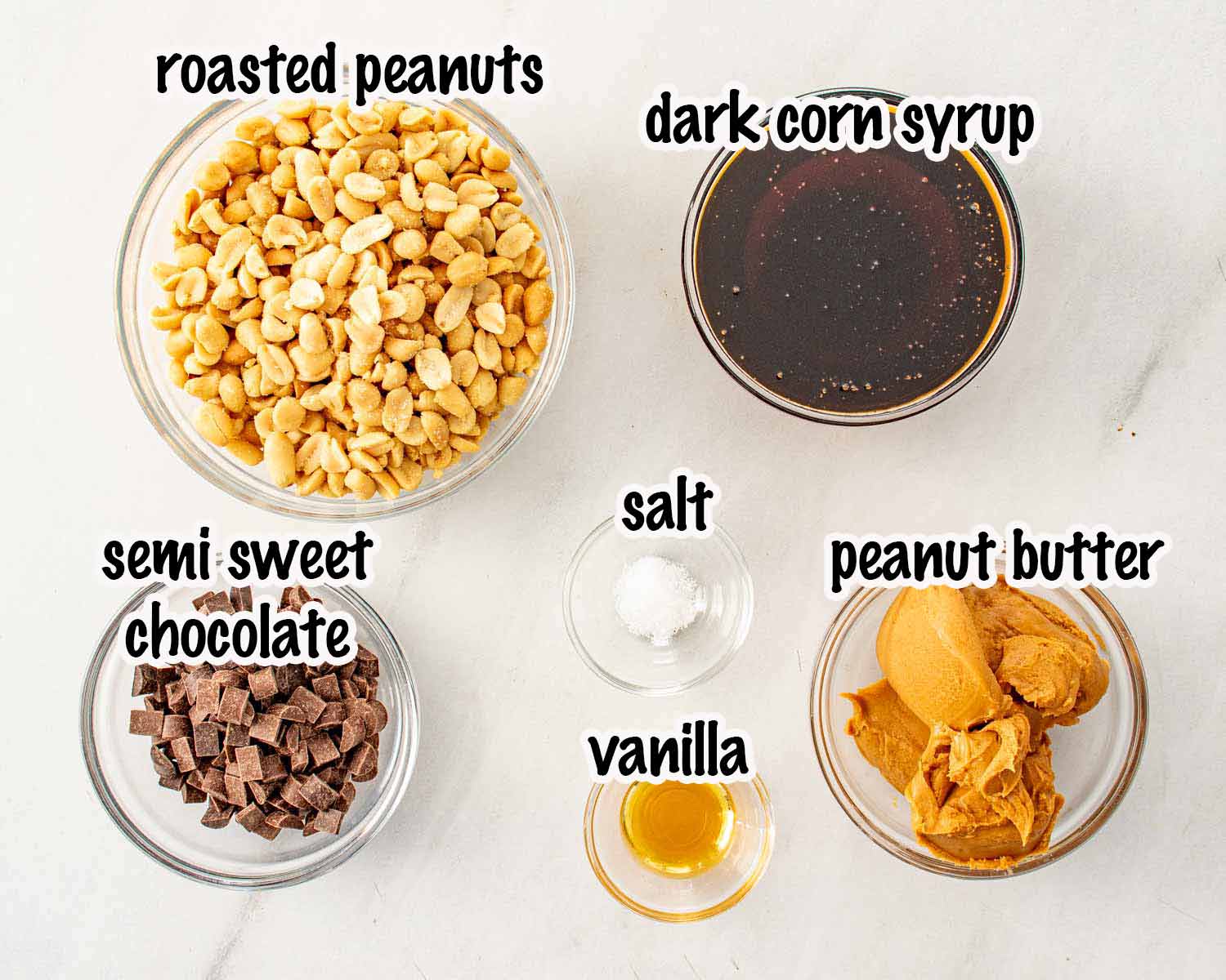 Glass bowls of roasted peanuts, chocolate, peanut butter, and syrup labeled in black text on a white counter.