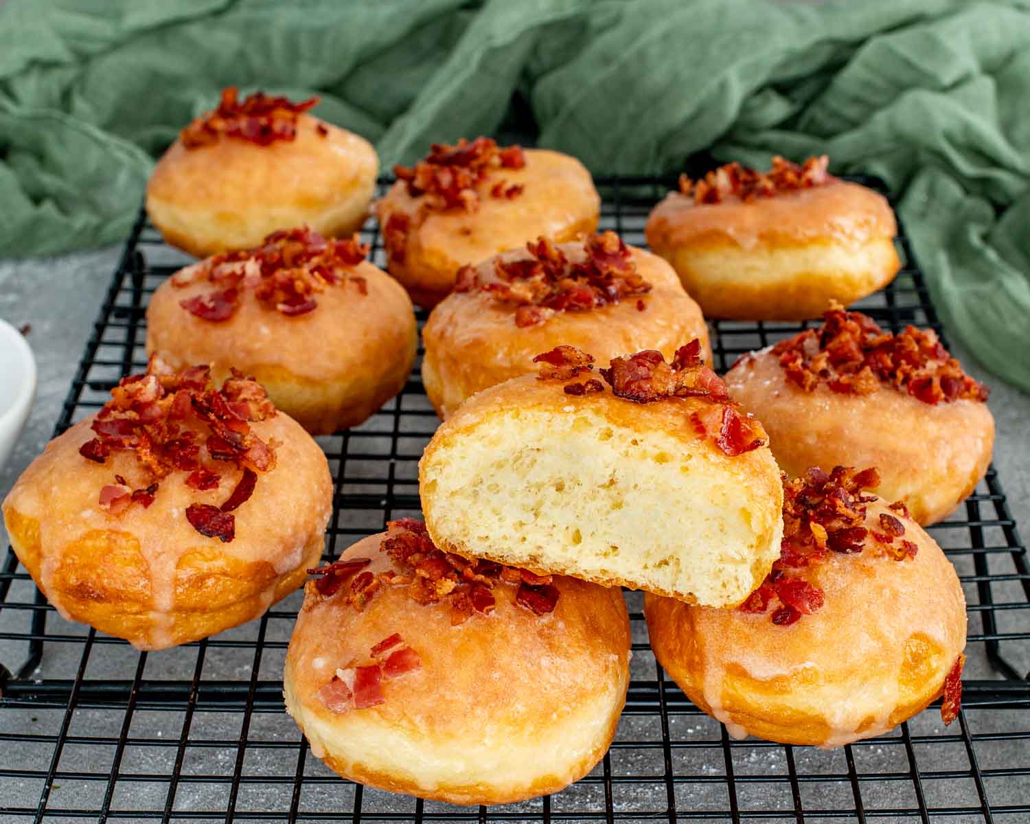 A row of glazed bacon donuts on a cooling rack with one donut sliced open to show airy interior.