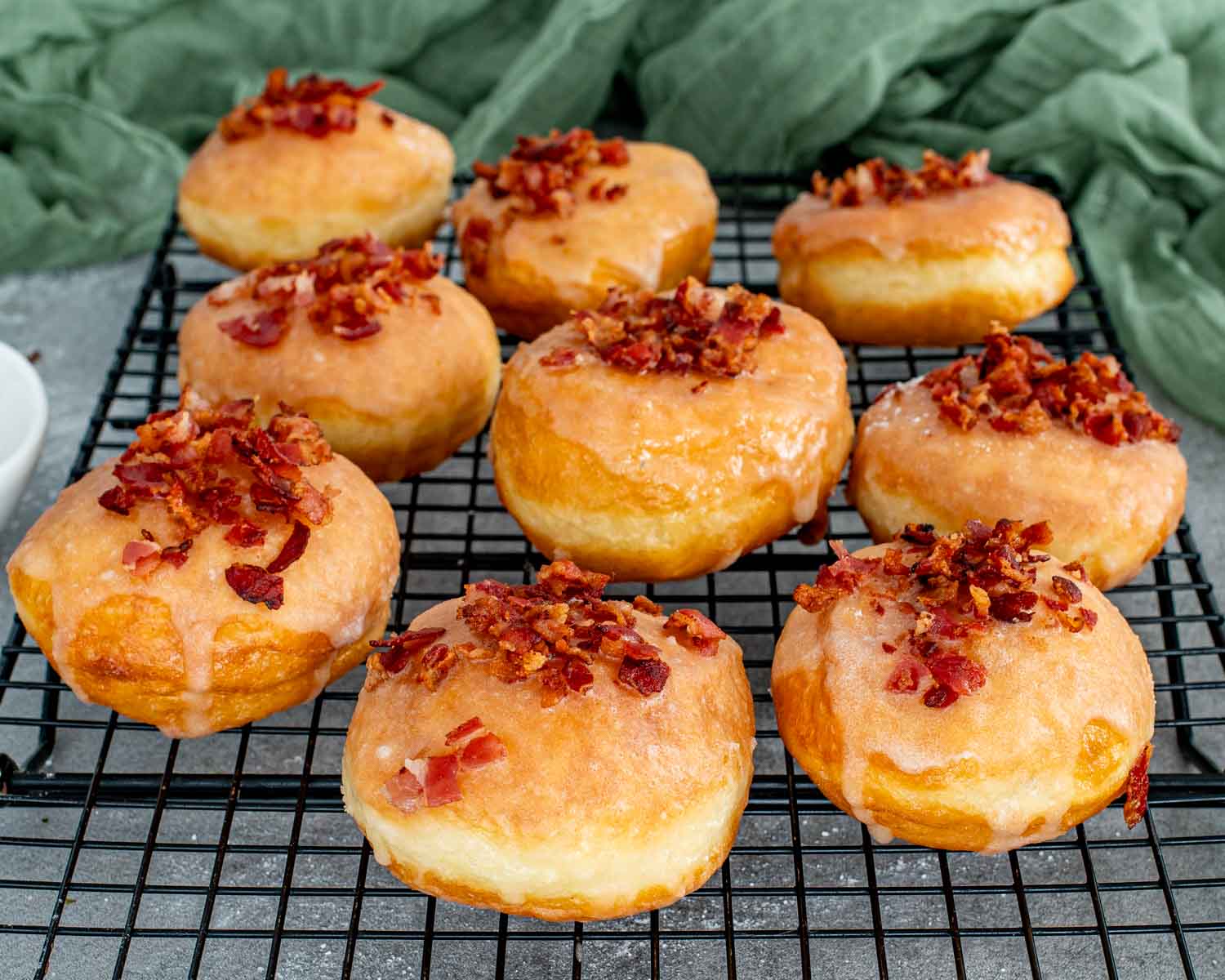 Nine perfectly fried donuts cooling on a wire rack topped with glossy maple glaze and sprinkles of crispy bacon.
