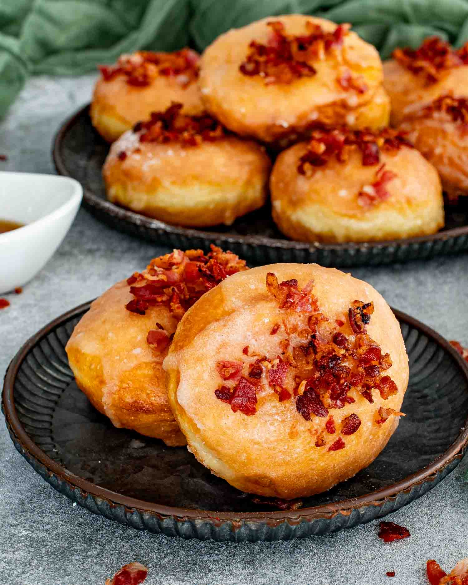 Two golden donuts sitting on a dark plate with a mountain of more glazed bacon donuts in the background.