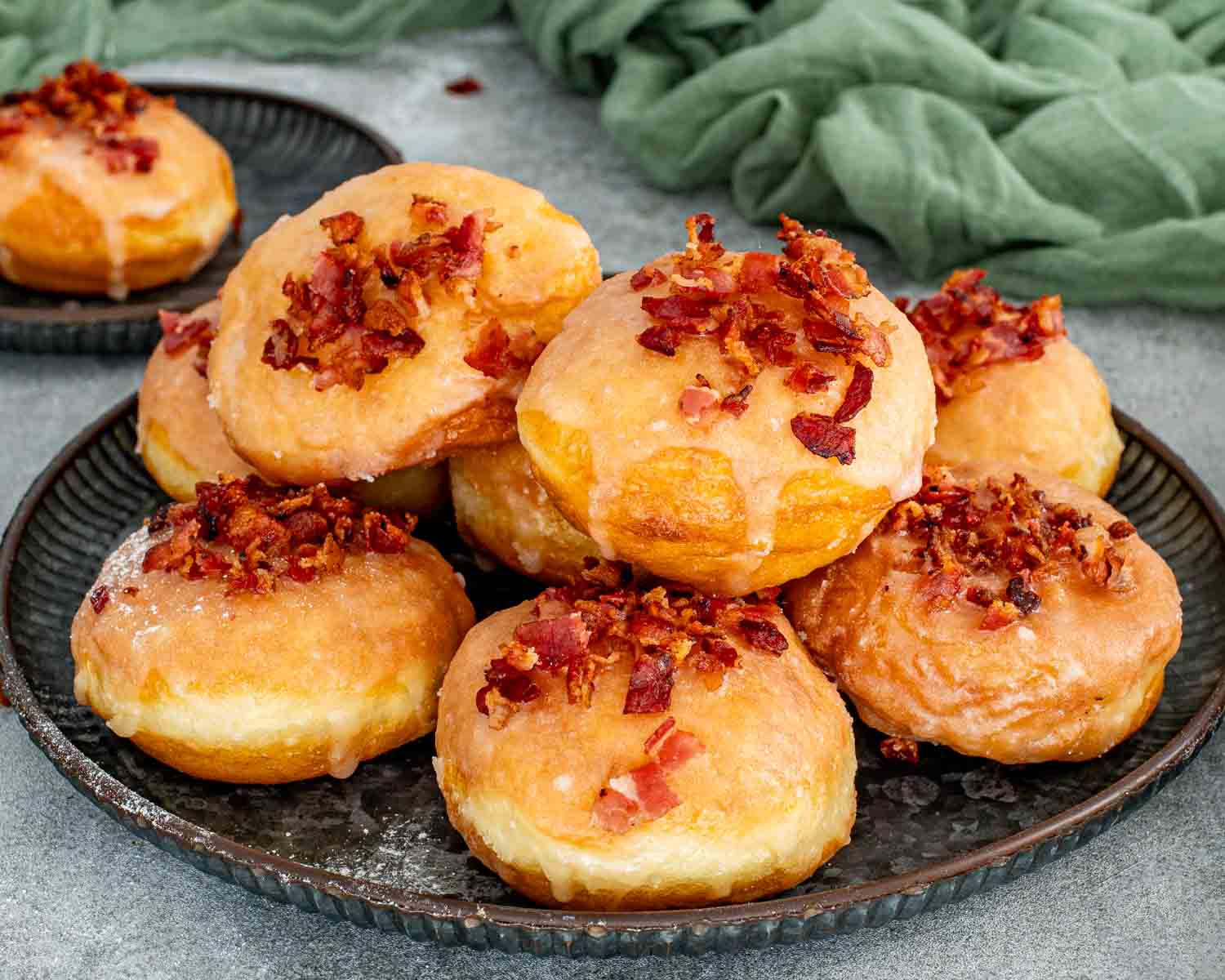 A large stack of golden brown donuts on a dark metal tray heavily topped with glaze and chopped bacon.