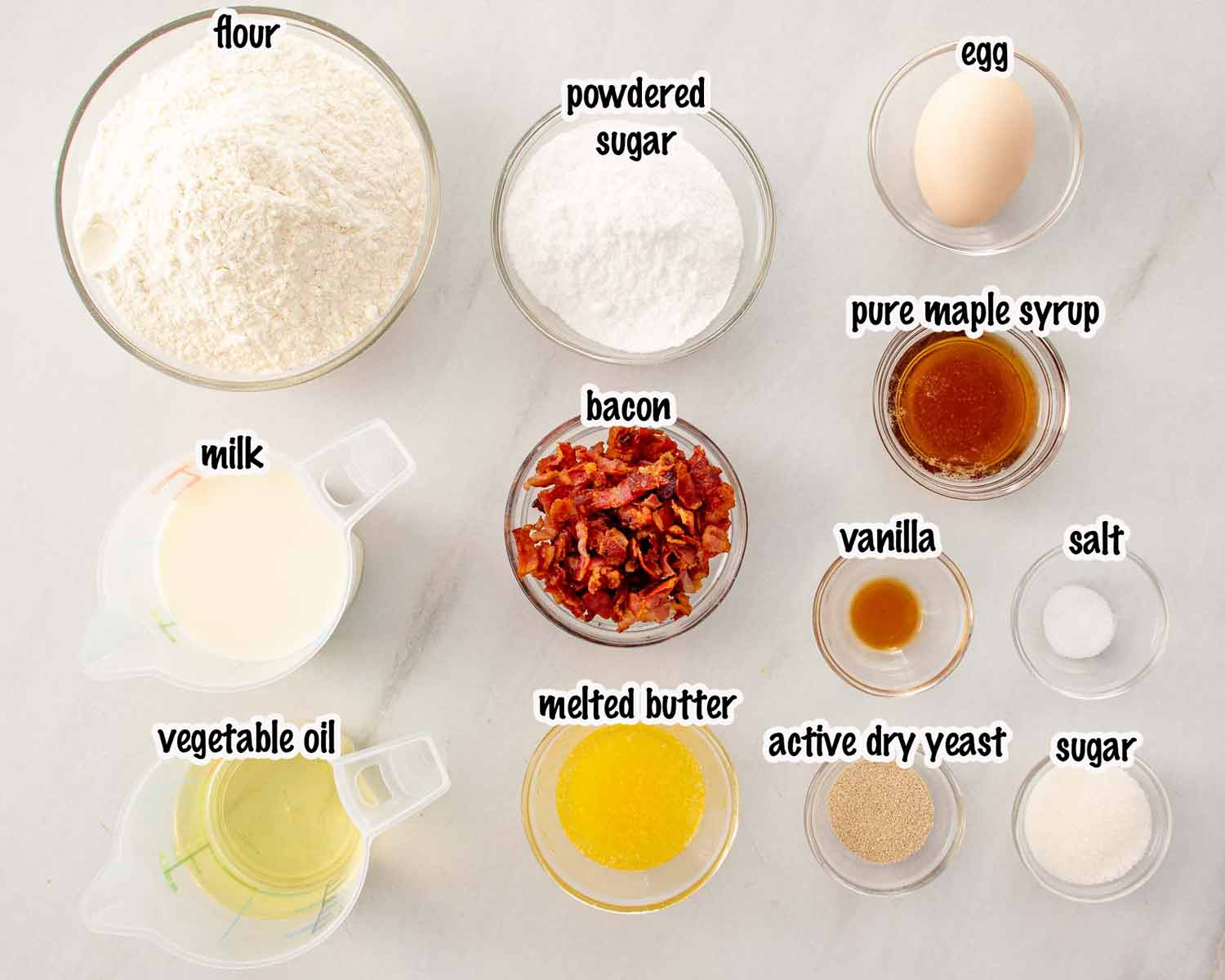 Top down view of bowls containing flour, sugar, yeast, bacon, maple syrup, milk, and eggs on a white marble surface.