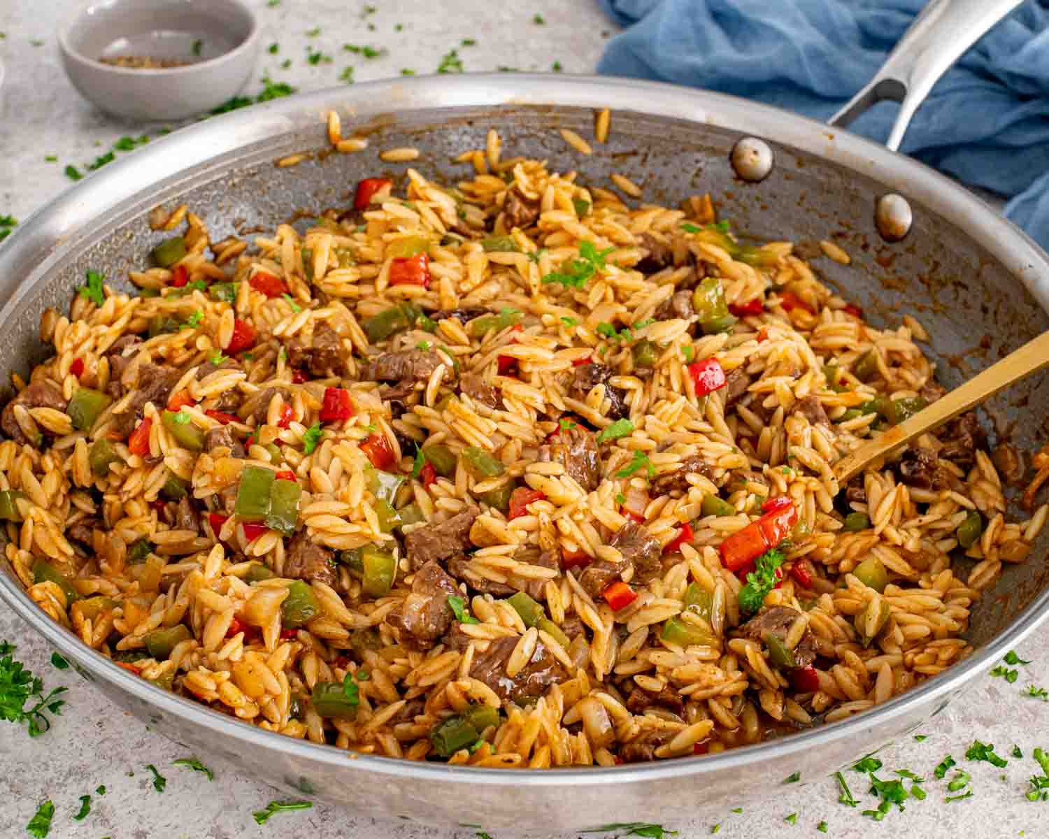 One pan beef and orzo pasta cooking in a stainless steel skillet with a gold spoon and fresh herb garnish.