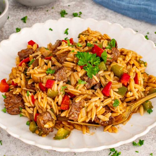 A white scalloped plate featuring a serving of beef and orzo pasta garnished with fresh parsley and a gold spoon.