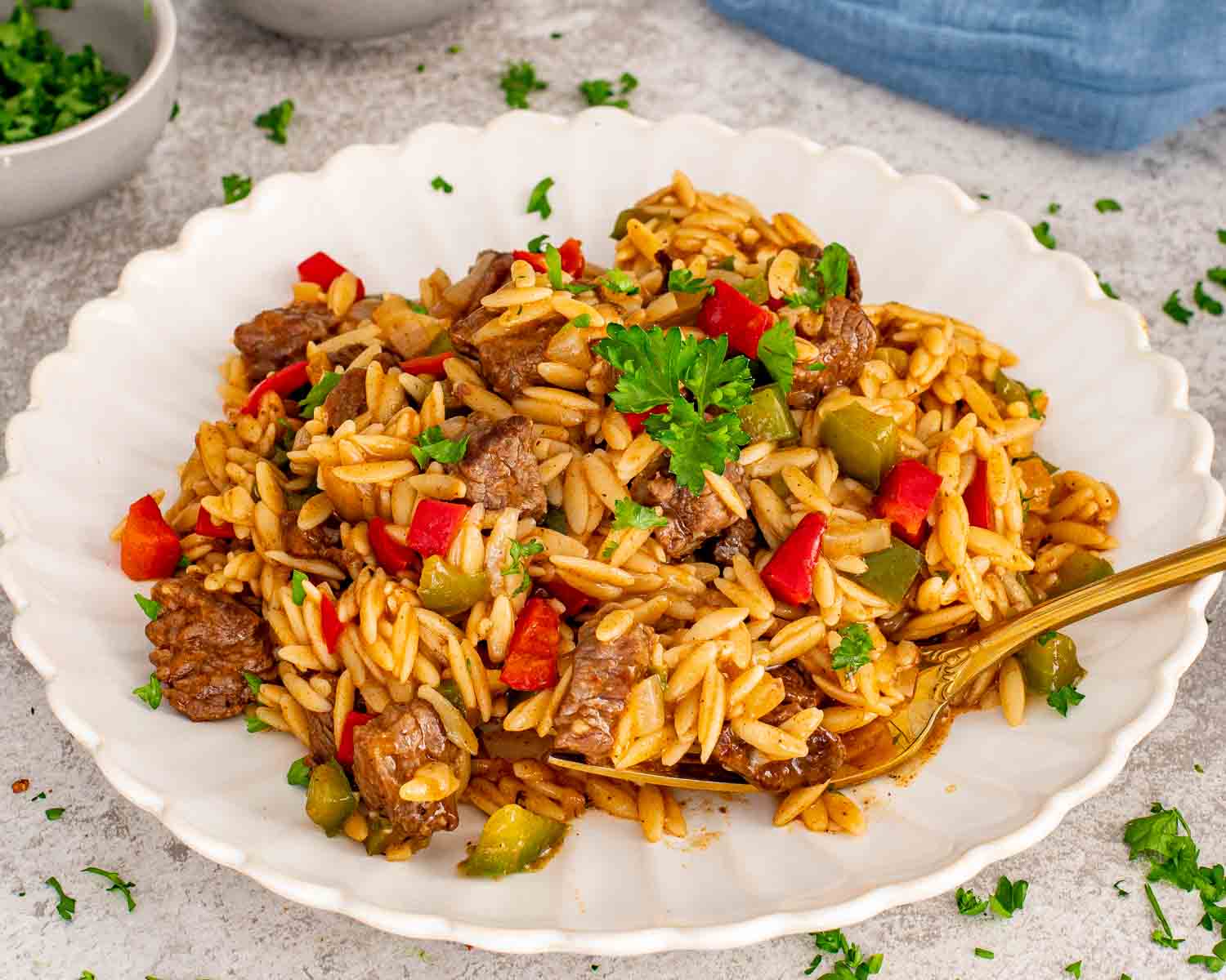 A white scalloped plate featuring a serving of beef and orzo pasta garnished with fresh parsley and a gold spoon.