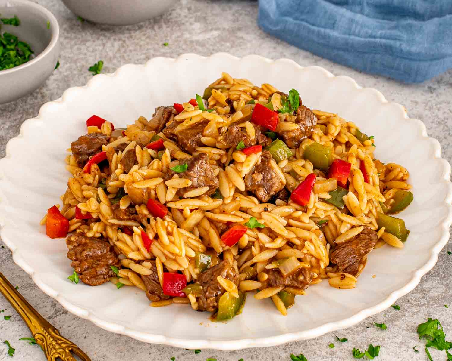 Close up shot of tender beef pieces and orzo pasta with peppers on a white plate with a blue napkin.