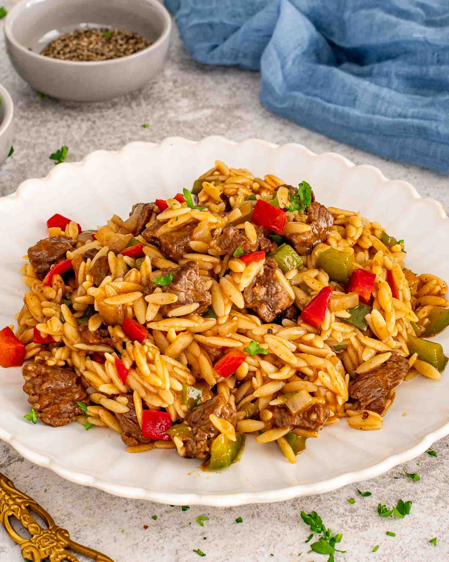 Close up shot of tender beef pieces and orzo pasta with peppers on a white plate with a blue napkin.